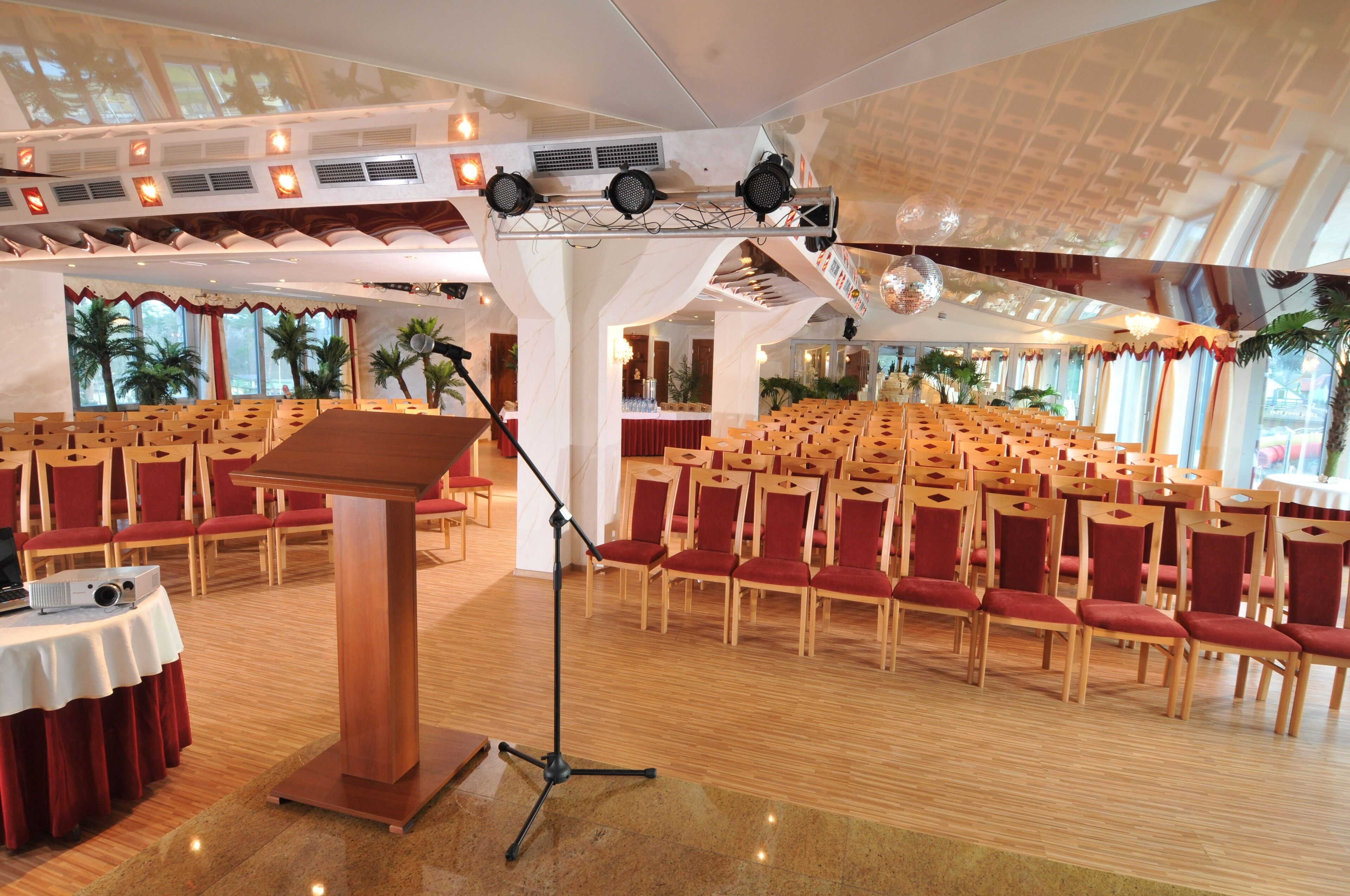 Un salón de conferencias con filas de sillas de madera y un atril, ventanas amplias con cortinas e iluminación en el techo.