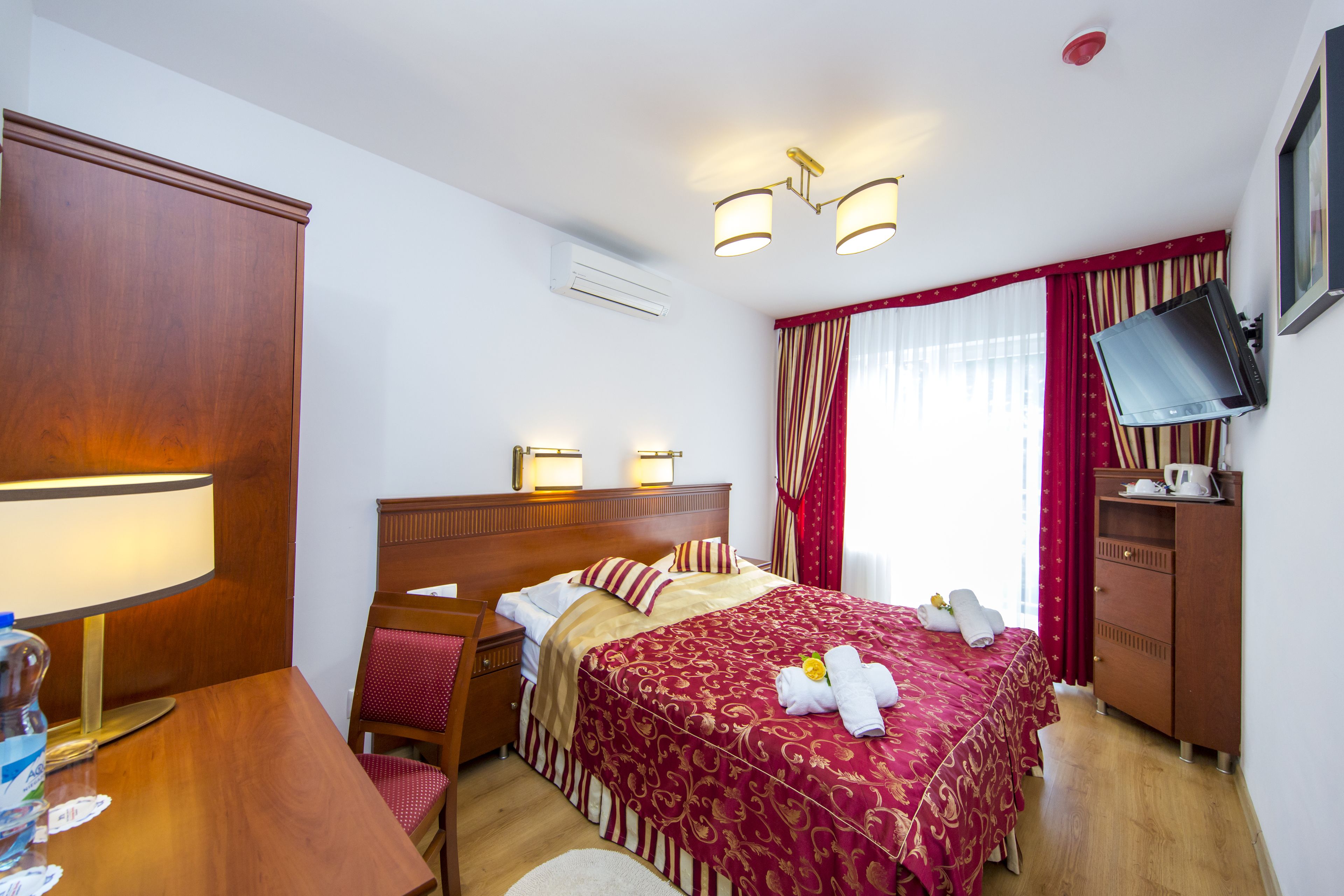 A hotel room with a wooden bed, two bedside lamps, a TV on a wooden cabinet, and a window with red curtains.