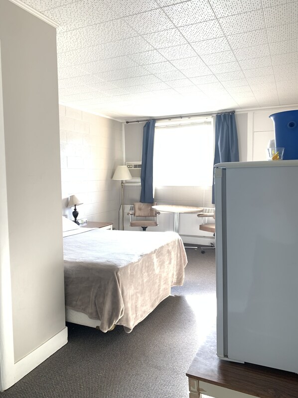 A hotel room with a bed, a desk, a chair, a lamp, a window with curtains, and a refrigerator.