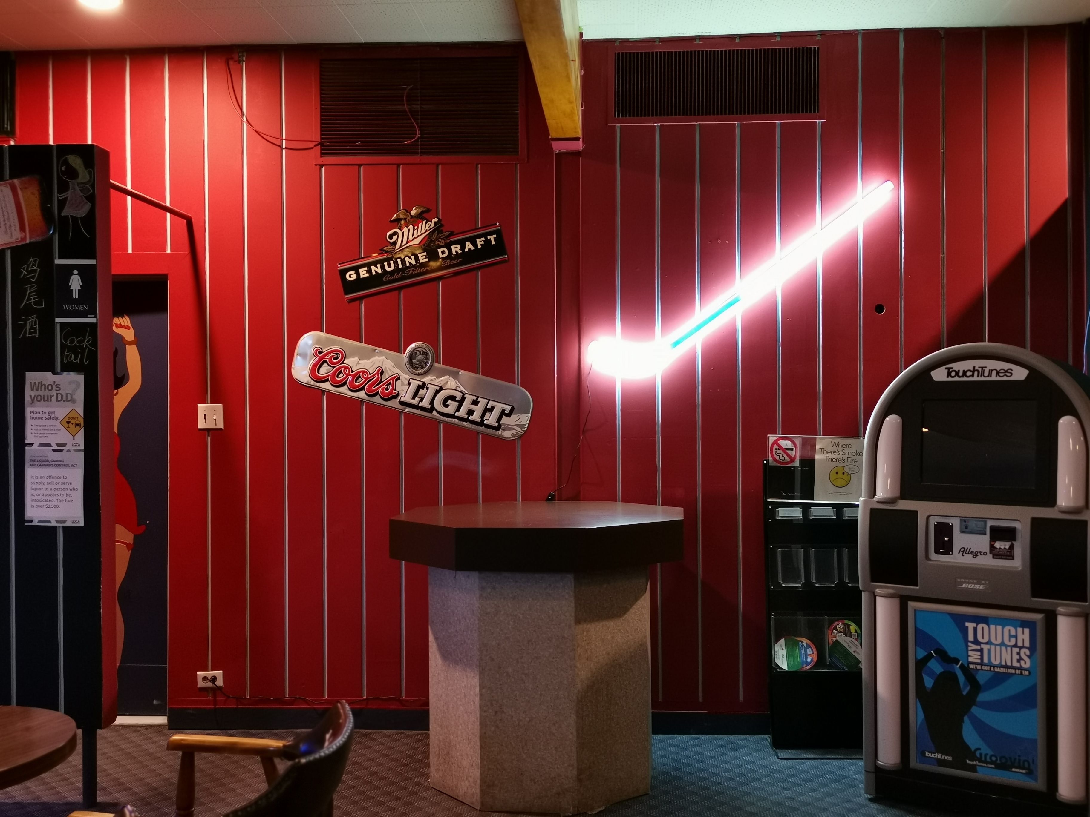 Un coin bar avec un mur rouge, une enseigne « Miller Genuine Draft », une enseigne « Coors Light » et un juke-box « TouchTunes ».