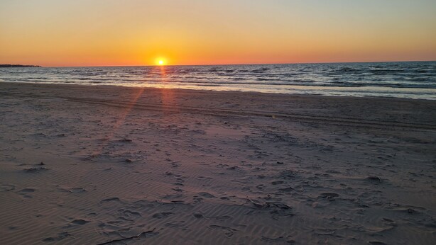 A beach with footprints leading to the water, a sunset in the background, and a calm sea. - The Pinewood - Adult Getaway by the Lake (Lambton Shores)