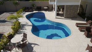 A curved swimming pool with a tiled pattern, surrounded by lounge chairs and a table. - Residencial Portinari (Porto Seguro)