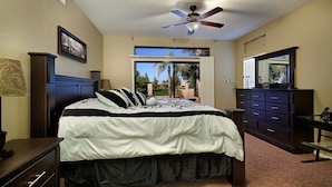 A bedroom with a bed, a dresser, a desk, a ceiling fan, and a window with a view of palm trees. - Chandler - 4 Bedroom Home - Chandler (Chandler)