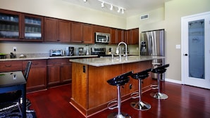 A modern kitchen with wooden cabinets, stainless steel appliances, a granite countertop, and a bar area with stools. - Chandler - 4 Bedroom Home - Chandler (Chandler)
