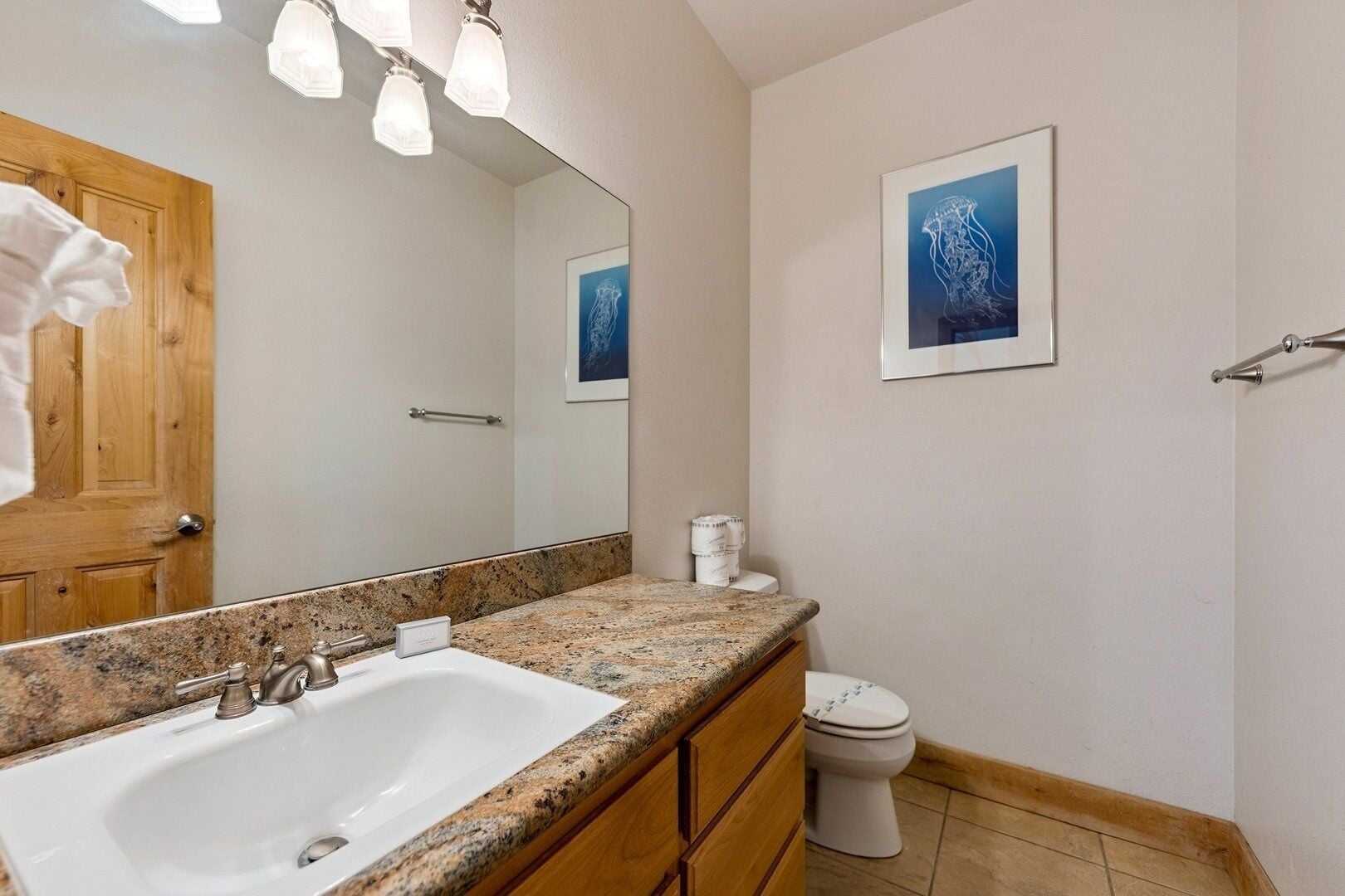 Ein Badezimmer mit Granitwaschtisch, Holzschränken, einer weißen Toilette und einem gerahmten Kunstwerk an der Wand.