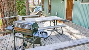 Una terraza de madera con una parrilla y una mesa de picnic, rodeada de árboles y un pequeño edificio.