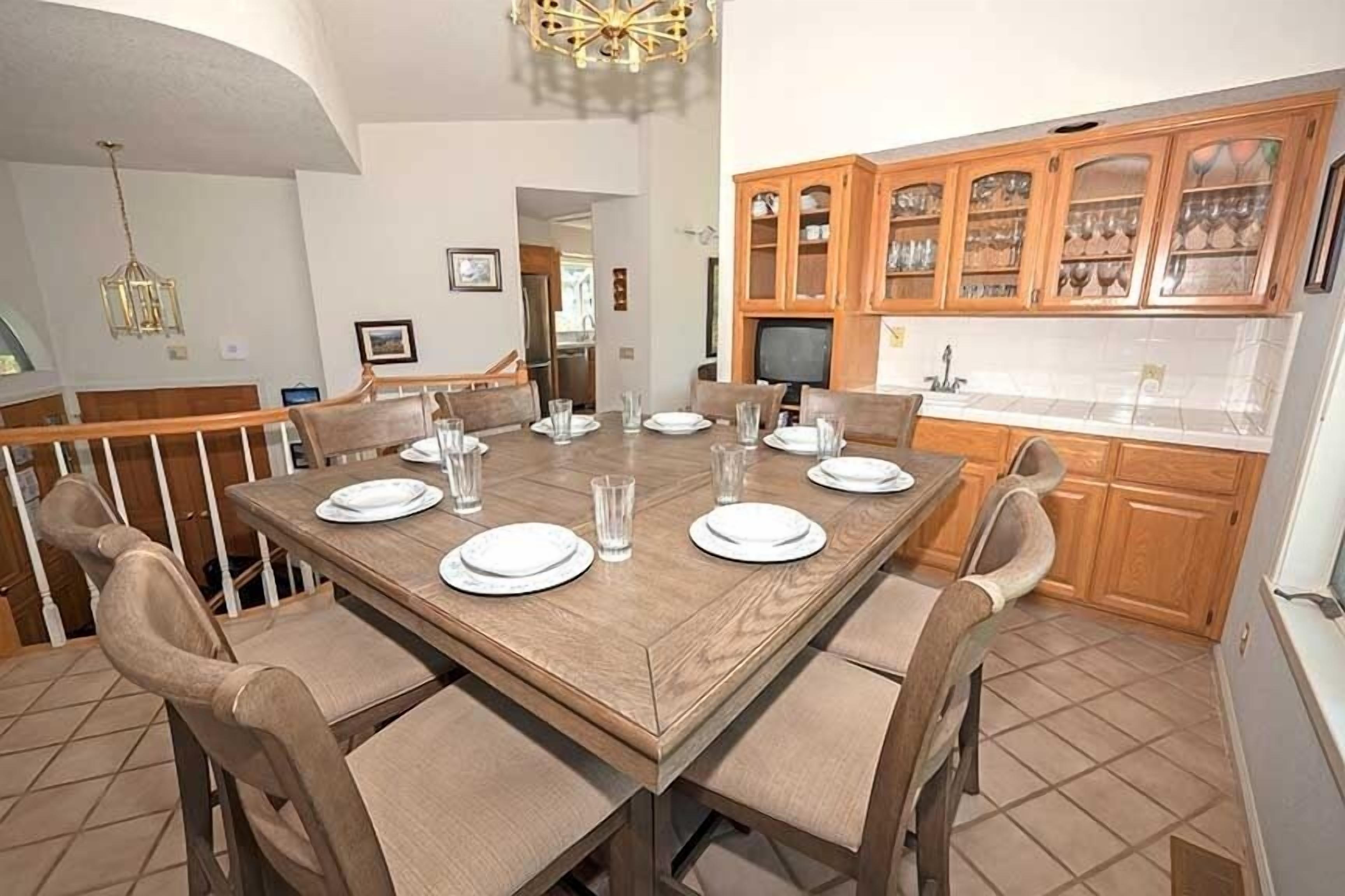 A dining area with a wooden table set for six, surrounded by cushioned chairs. A television is mounted on the wall above the table.