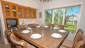 Un comedor con una mesa de madera dispuesta para seis personas, rodeada de sillas de madera. Hay una zona de cocina con armarios de madera y un televisor a la izquierda.
