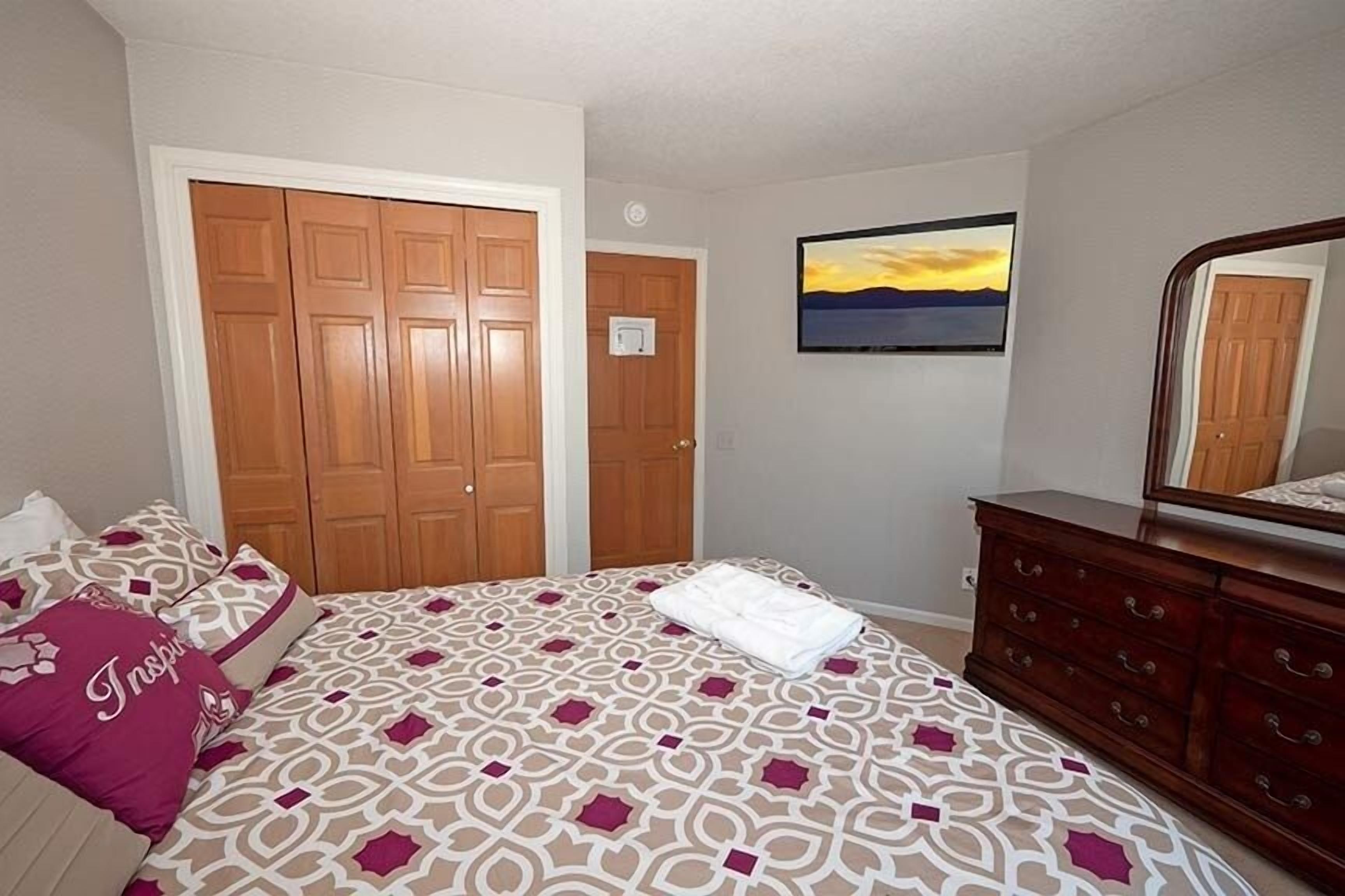 A bedroom with a patterned bedspread, a wooden wardrobe, a dresser, and a painting of a sunset.