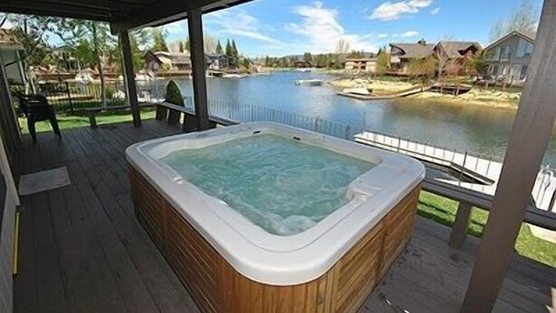 Un bain à remous sur une terrasse en bois, donnant sur un lac avec des maisons en arrière-plan.