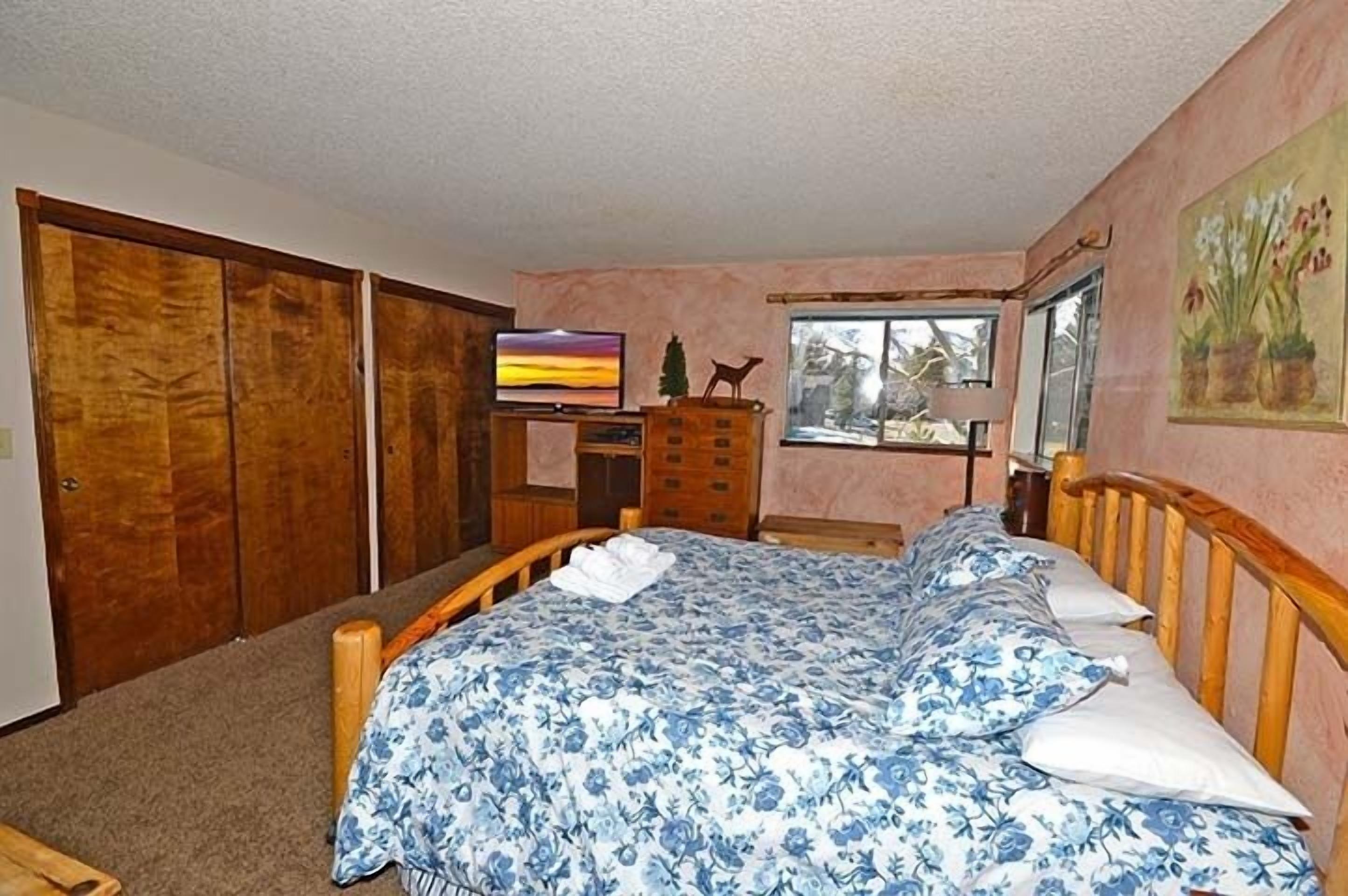 Un dormitorio con cama, tocador, televisor y ventana con vista a árboles.