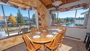 Un coin repas avec une table et des chaises en bois, de grandes fenêtres offrant une vue sur les montagnes et un lac, et un panneau indiquant les locations.