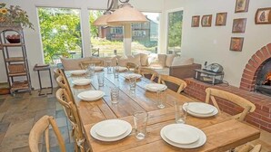 A dining area with a long wooden table set for six, surrounded by wooden chairs. There is a brick fireplace and a large window providing a view of greenery outside. - 2130 Monterey Drive 4 Bedroom Home by RedAwning (South Lake Tahoe)