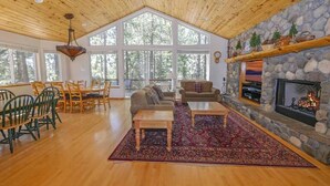 A spacious living room with a stone fireplace, wooden ceiling, and large windows. - 2460 Lupine Trail 6 Bedroom Home by RedAwning (South Lake Tahoe)