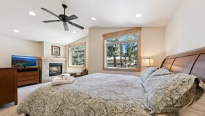 A bedroom with a large bed, a ceiling fan, a fireplace, a TV, and a window with a view of trees.