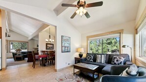 A spacious living area with a sofa, coffee table, and a dining table with chairs. There is a ceiling fan, a painting on the wall, and a window providing natural light.