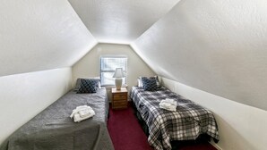 A small, triangular attic bedroom with two single beds, a nightstand, a lamp, and a window with blinds.
