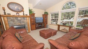 A living room with a stone fireplace, leather sofas, a television, and large windows with a view of greenery.