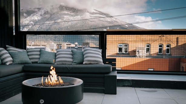 A modern outdoor seating area with a fire pit, a sofa with striped cushions, and a view of a mountain and buildings through a wire mesh. - Eleven Revelstoke Lodge (Revelstoke)