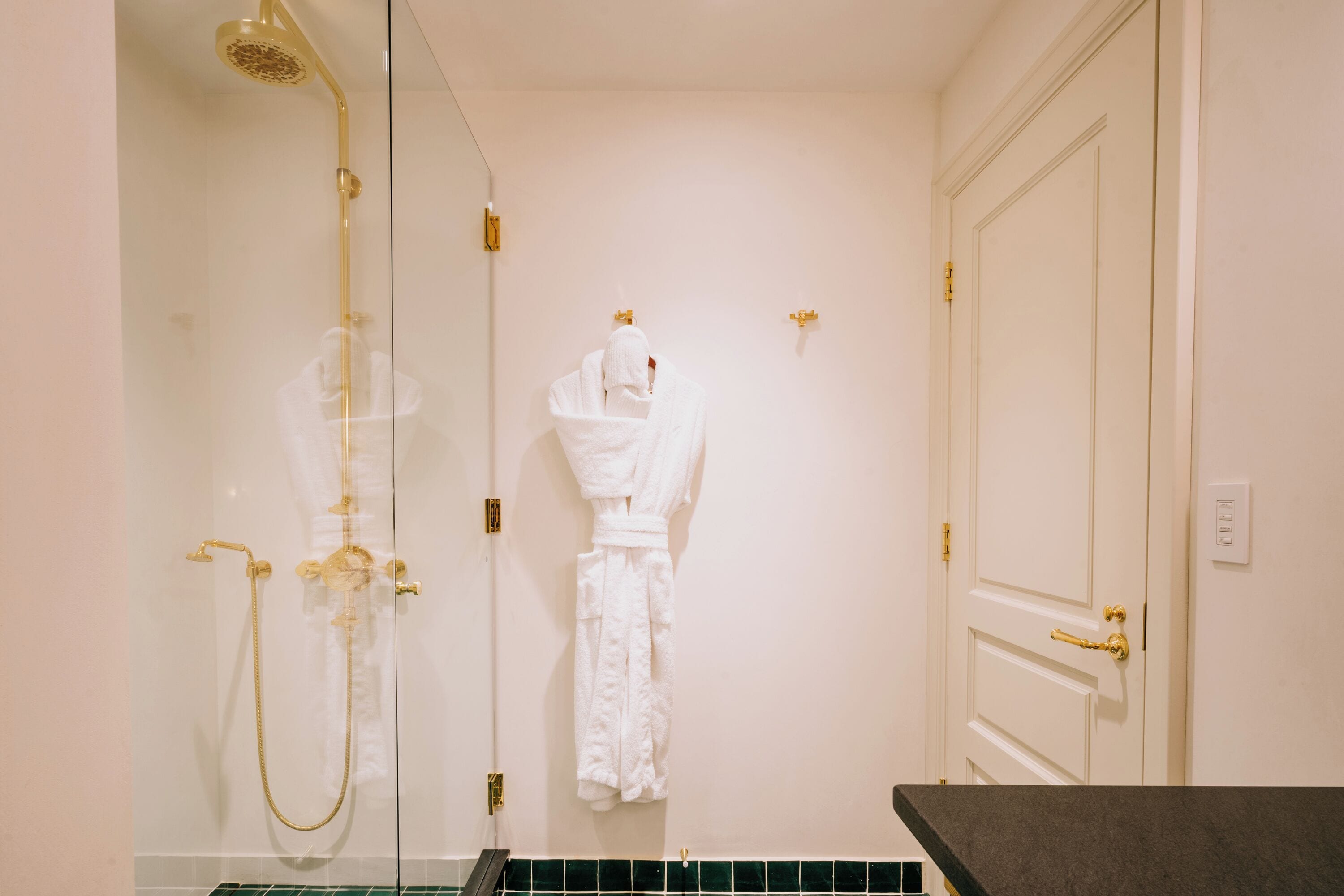 A bathroom with a glass-enclosed shower, a white robe hanging on the wall, and a white door with a gold handle.