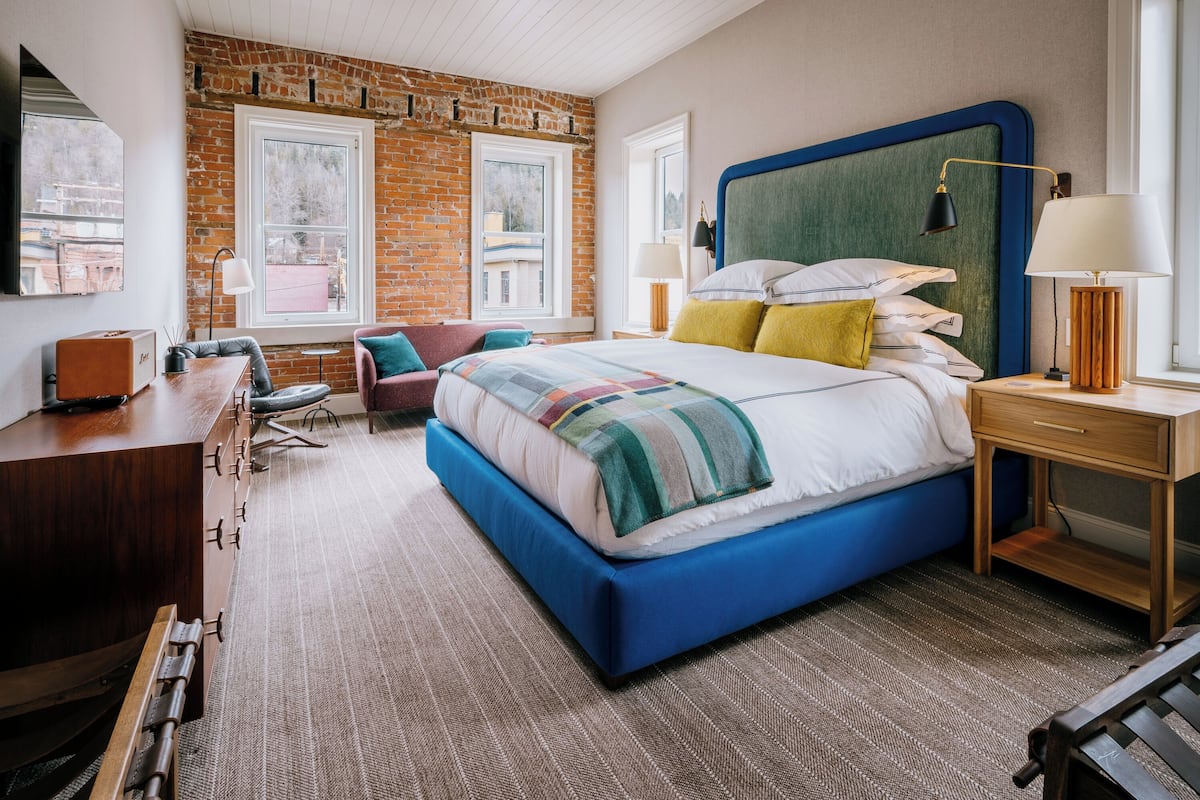 Une chambre d’hôtel moderne avec un grand lit, un bureau et une chaise. Elle comporte deux fenêtres et un mur en briques.