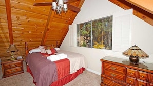 A bedroom with a sloped ceiling, a bed with red bedding, a wooden dresser, and a window with a view of trees. - 3542 Bobby Grey Circle 3 Bedroom Cabin by RedAwning (South Lake Tahoe)
