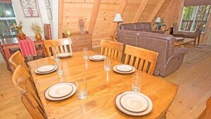 Una mesa de comedor de madera dispuesta con platos y vasos, rodeada de sillas de madera en una habitación acogedora al estilo de una cabaña de troncos, con un sofá y una ventana.