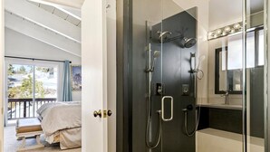 Une salle de bain moderne avec une douche à l’italienne en verre, un grand miroir et une vue sur un balcon aménagé avec un lit.