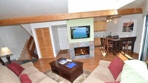 A living room with a fireplace, a TV mounted on the wall, and a dining area with chairs and a table.
