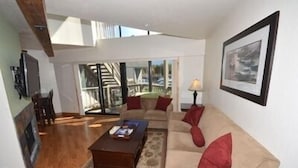 A living room with a sofa, coffee table, and a TV. There is a staircase leading to a balcony.