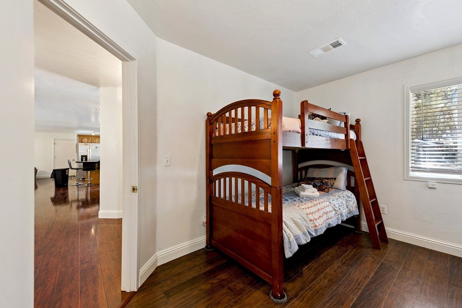 A bedroom with a wooden bunk bed, a window with blinds, and hardwood flooring.
