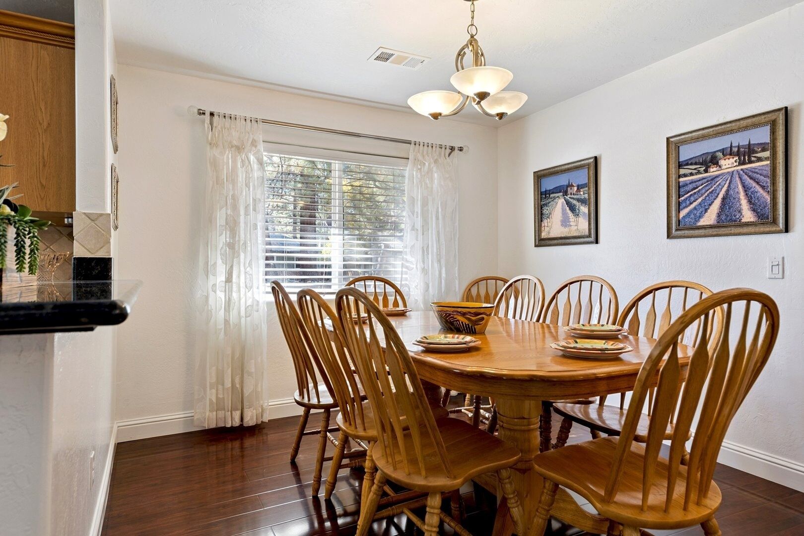 A dining room with a round wooden table, six wooden chairs, and two framed paintings on the wall.