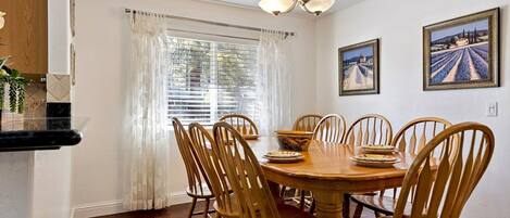 A dining room with a round wooden table, six wooden chairs, and two framed paintings on the wall.