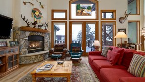A cozy living room with a stone fireplace, a red sofa, and a large window overlooking a snowy deck. - Crosstimbers 2784 3 Bedroom Townhouse by RedAwning (Steamboat Springs)