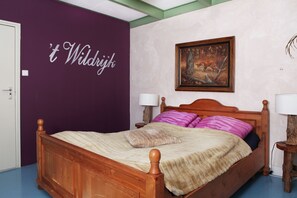 A bedroom with a wooden bed, purple walls, a framed painting, and a 't Wildrijk' sign.