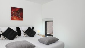 A hotel room with a large bed, two black and one grey pillow, a white headboard, a red abstract painting on the wall, a window with a view of a brick building, and a bedside lamp.