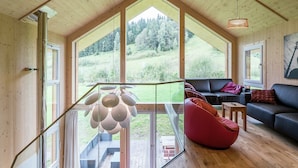 Un loft spacieux avec un plafond haut, de grandes fenêtres offrant une vue sur un paysage verdoyant, un lustre moderne et un coin salon confortable avec un canapé et un fauteuil poire.