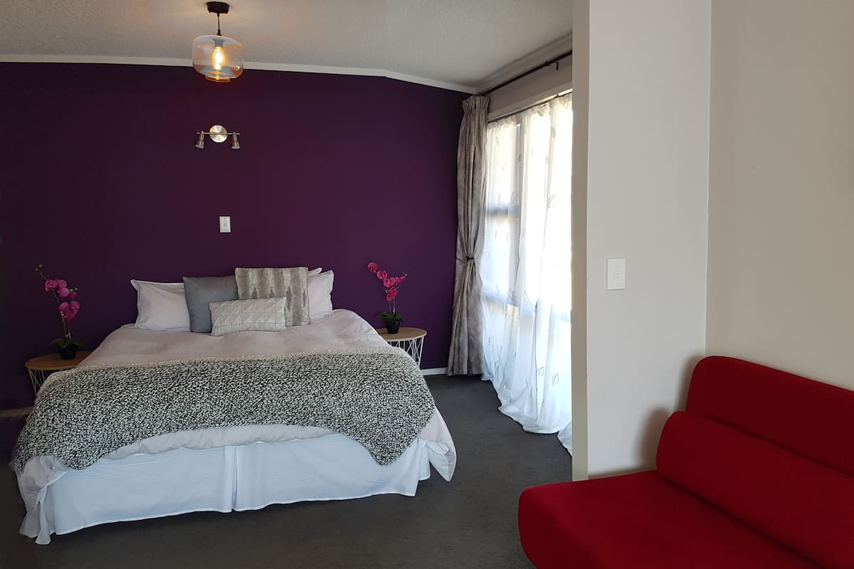 A bedroom with a bed, a red chair, a small table, and a window with curtains.