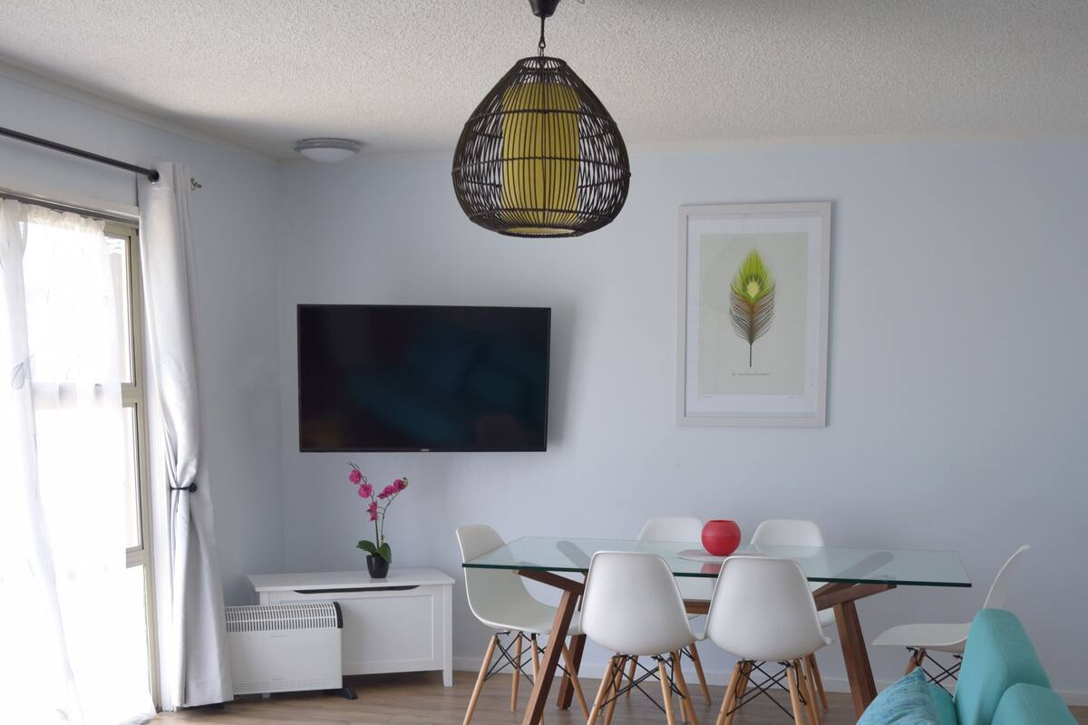 A modern living room with a teal sofa, a glass coffee table, a dining area with a glass table and white chairs, a flat-screen TV mounted on the wall, and a hanging light fixture.