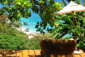 A wicker chair with a table and an umbrella, overlooking a beach with clear blue water and lush greenery. - Beijupira Lodge Noronha Pousada (Fernando de Noronha)