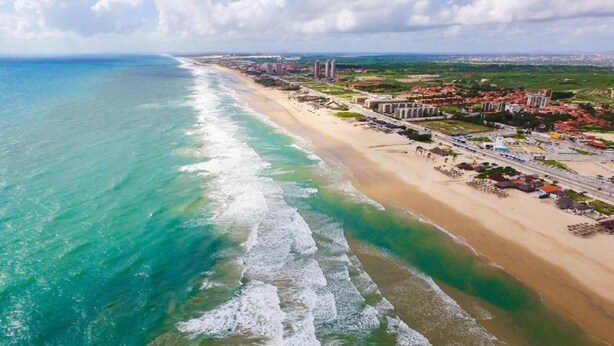 Luchtbeeld van een strand met helder turquoise water, een zandstrand en een stadsbeeld met gebouwen en wegen op de achtergrond.