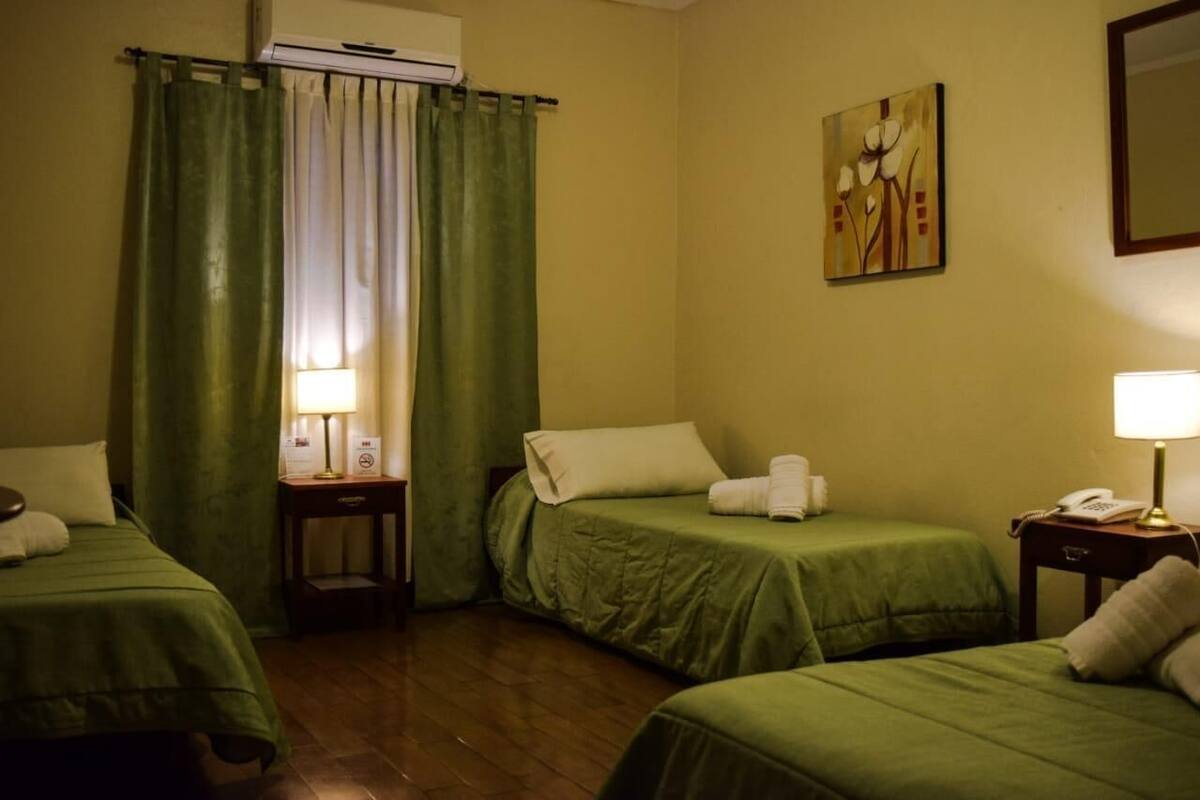 A hotel room with two single beds, a green curtain, a painting on the wall, and an air conditioning unit.