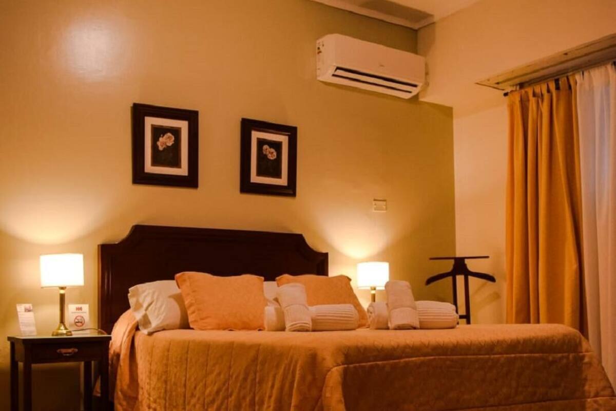 A hotel room with a large bed, two bedside lamps, an air conditioning unit, and a window with curtains.