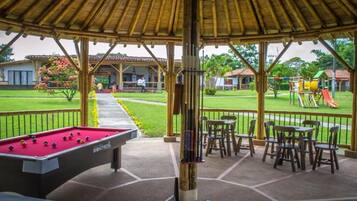 Una mesa de billar bajo una zona techada, con asientos al aire libre y un parque infantil al fondo.