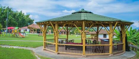 A wooden gazebo with a green roof, surrounded by a grassy area and playground equipment.