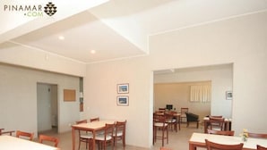 A spacious dining area with multiple wooden tables and chairs, a small kitchen area, and framed pictures on the walls. - Portasol Apart Hotel (Pinamar)