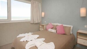 A bedroom with a bed, pillows, a nightstand, a lamp, and a window with a view. - Portasol Apart Hotel (Pinamar)