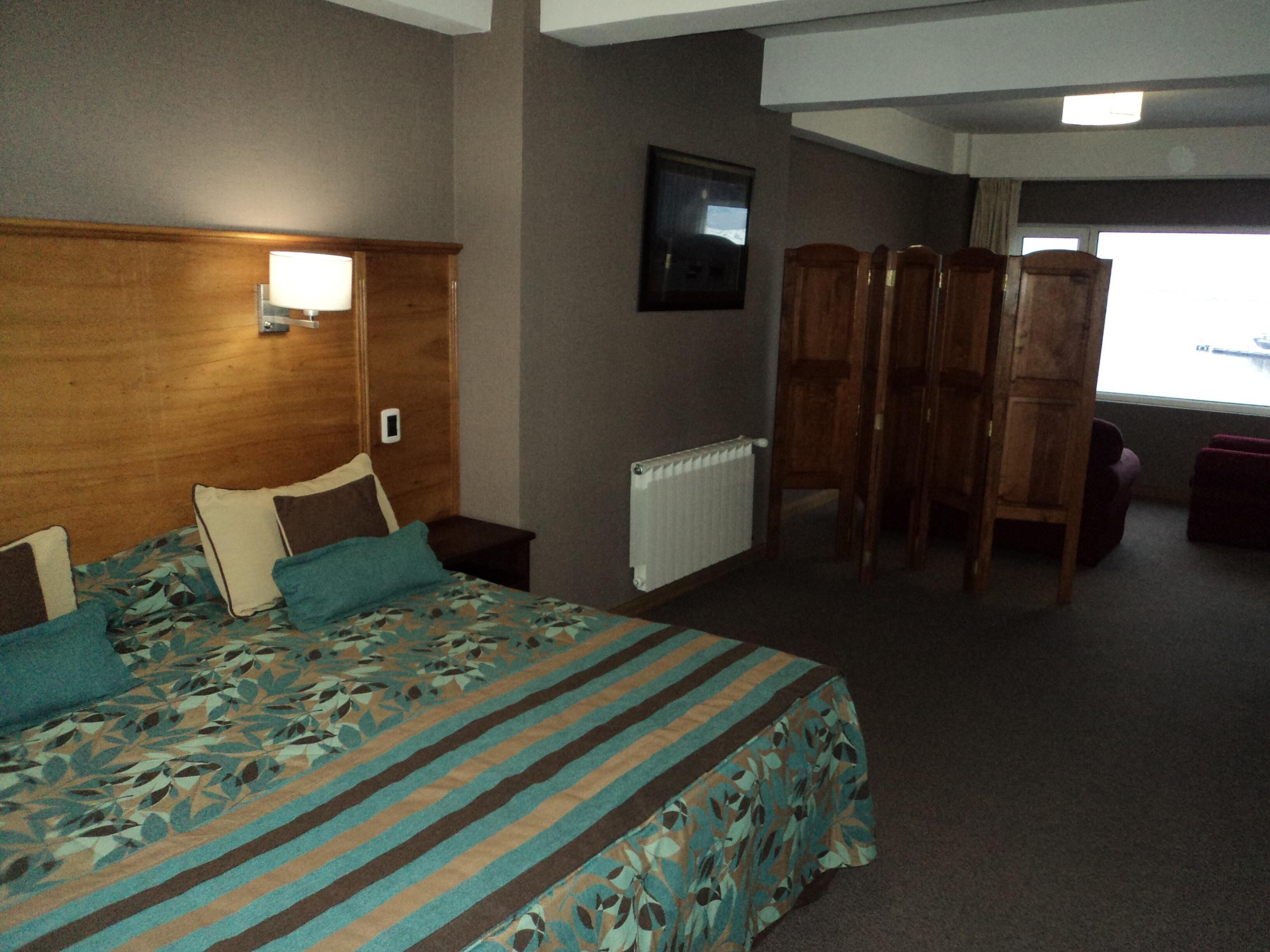 Habitación de hotel con cama, cabecera de madera, televisor montado en la pared, radiador y ventana con vistas.