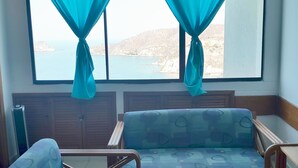 A room with two blue patterned chairs, a window with teal curtains, and a view of a body of water and hills. - Apartamento Tropical - Smr245a (Santa Marta)