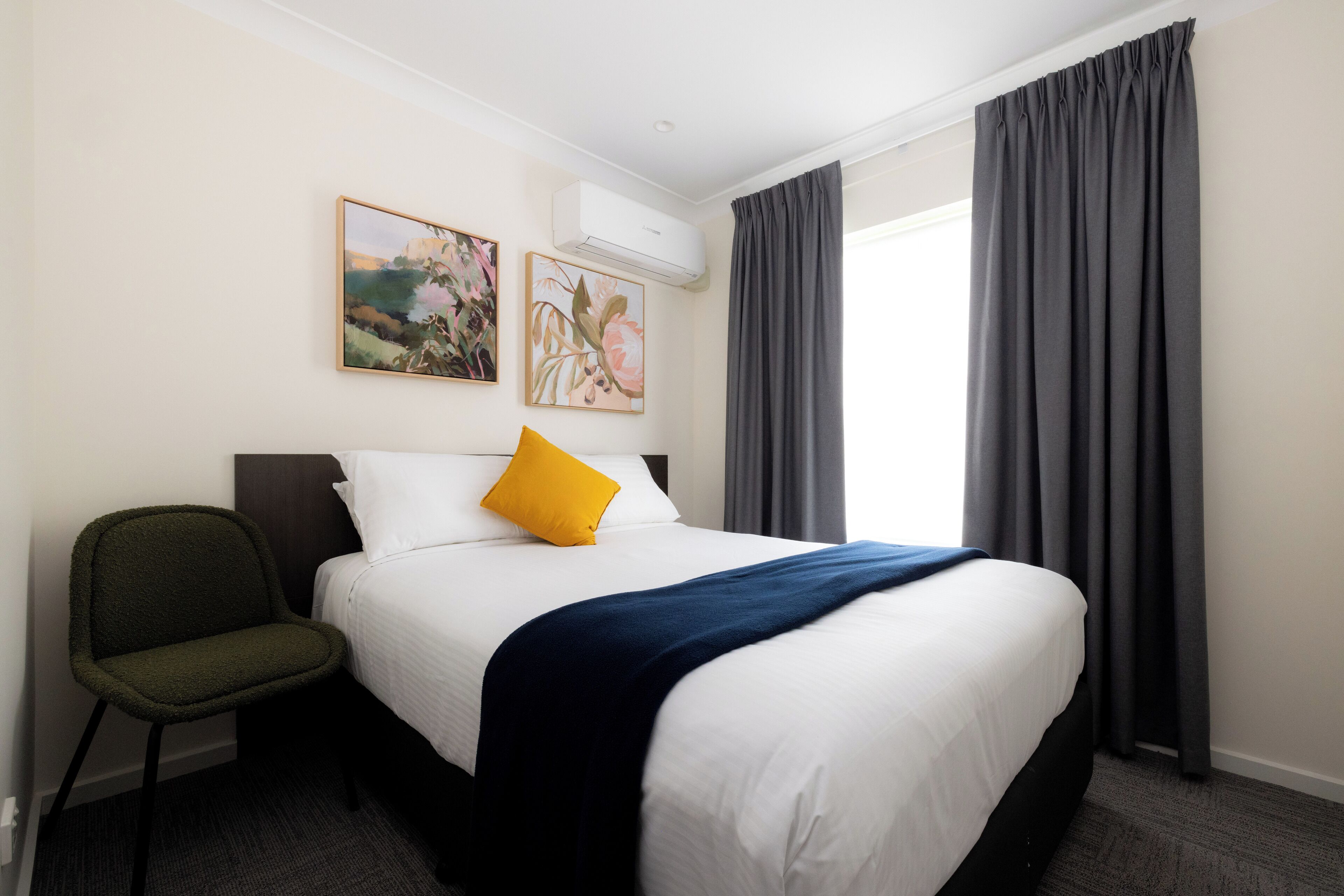 Een hotelkamer met een bed, een stoel, twee schilderijen aan de muur en een raam met gordijnen.
